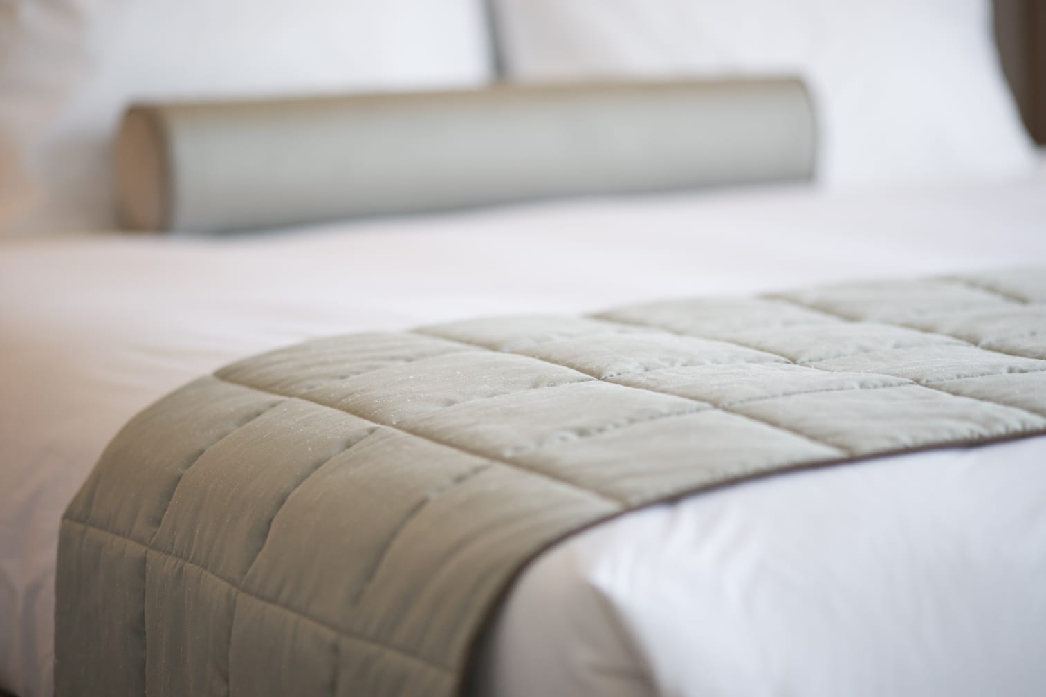 A neatly made bed with a quilted cooling mattress pad in a soft, neutral color. The bed is adorned with white linens and a cylindrical bolster pillow, creating a clean and inviting look.